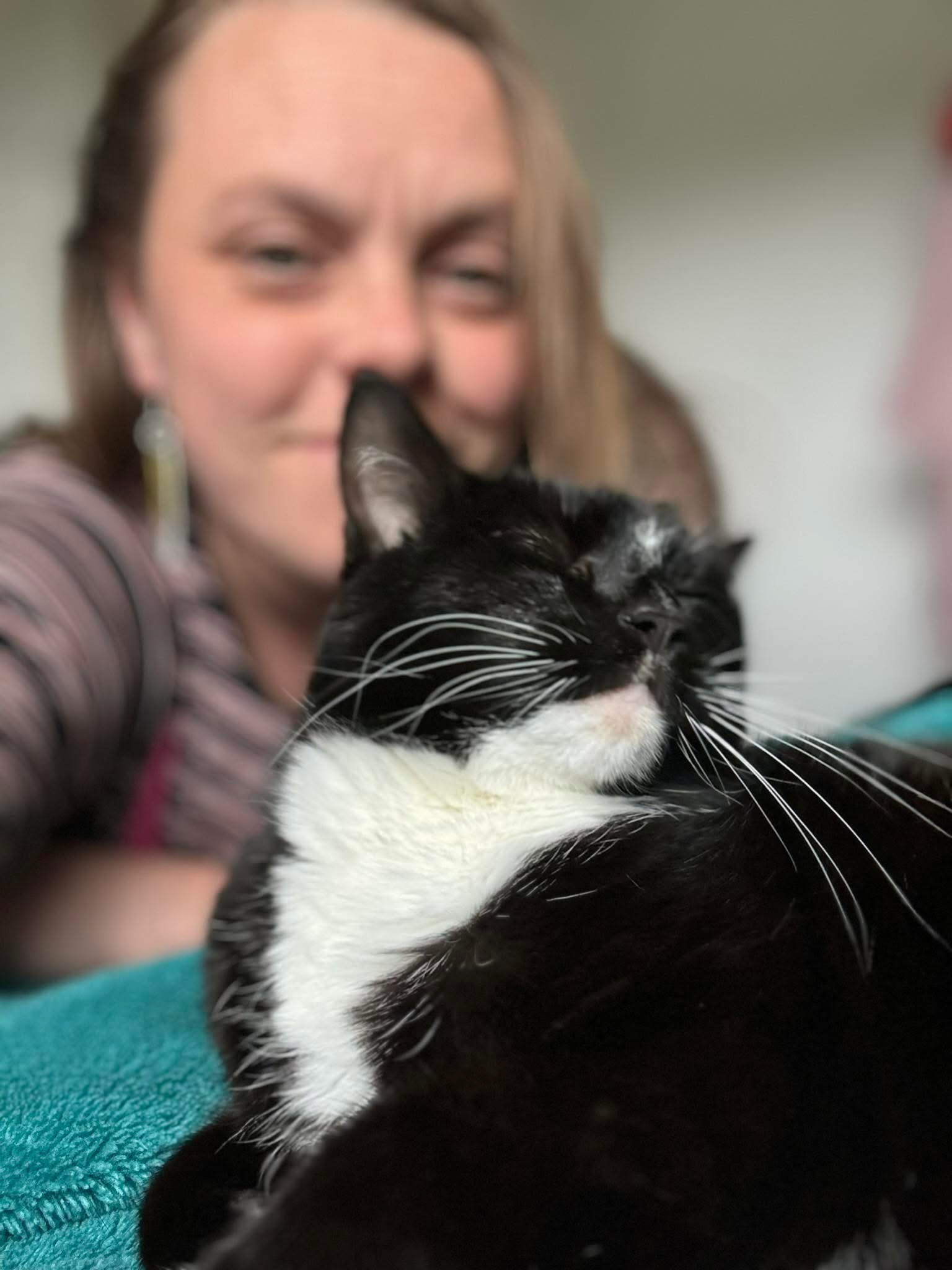 Leah smiling gently behind her own tuxedo cat Clara, who is purring with eyes closed against her shoulder